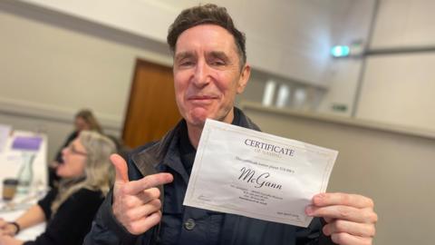 Paul McGann is smiling and holding a certificate of naming for a planet called McGann, his co-star Daphne Ashbrook is sat behind him and out of focus