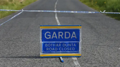 A stock photo of a Garda (Irish police) sign in the middle of a road.  The blue and white sign says "road closed" in both Irish and English.  A blue and white tape cordon stretches across the road behind the sign. 