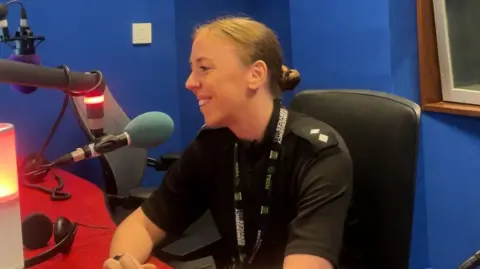Nicola Davis-Lyons, wearing a black police uniform, sits in a black chair in a radio studio speaking into a blue microphone.