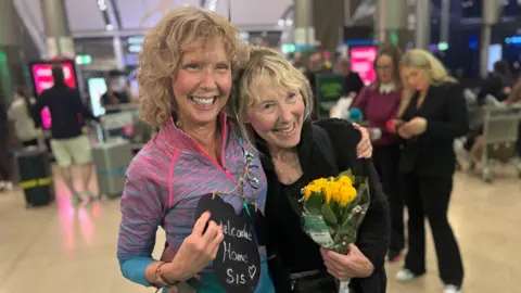 Elaine and Norita standing in an airport terminal, arms around each other and broad smiles on their faces - one is holding a "welcome home sis" sign and the other is holding a bunch of yellow roses
