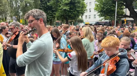 In the foreground, a man plays the flute. Behind him is a young boy concentrating as he plays the fiddle. In the background hundreds of other musicians play their instruments.