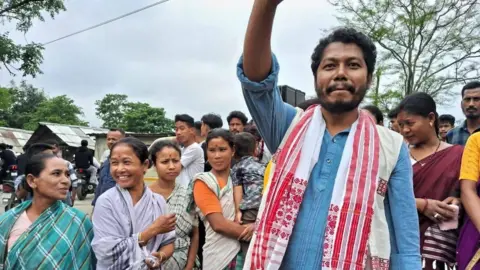 Youth for Bokakhat Pranab Doley - seen in a blue tunic with a red-white gamusa - raises his hand as he stands around a group of women in Assam