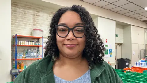Sarah Edwards is pictured with a closed mouth smile, wearing large circular black framed glasses, a green fleece jacket and blue jumper. She has black tight curly hair. In the background are green boxes and a shelf with various drinks bottles on it.