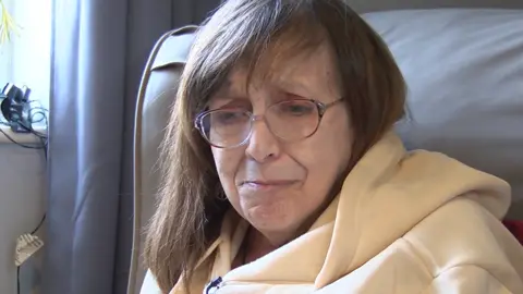 Lisa Barrett has brown hair and glasses and is wearing a hooded top in yellow and looks close to tears