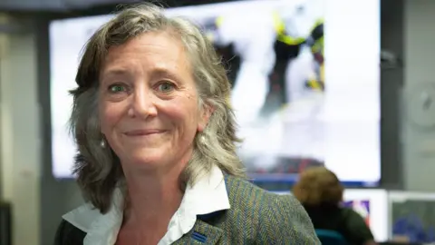 BBC News A smiling woman looks at the camera. She is wearing a white short and grey blazer. Behind her is an office featuring a large screen showing a map