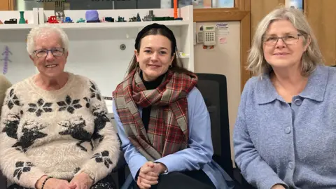 Three women are sat down looking at the camera and smiling. The women on the left has short grey hair and a beige and black Christmas jumper on. The women in the middle has long brown hair and a large tartan scarf, she is also wearing a black headband. The women on the far right is wearing a blue jumper and has short fair hair. 