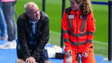 Eddie Mitchell DJ Fatboy Slim wearing a black leather jacket on his knees delilvering CPR to a dummy with an air ambulance professional next to him. She is wearing orange overalls 