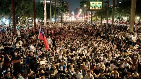 Image of Getty is shooting at night with thousands of protesters sitting on a boulevard in Taipei, holding up protest signs. In the foreground you can see a large Taiwan flag, while in the distance you can see the bright city lights of Taipei