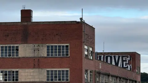 A derelict factory building. There is graffiti on one of the upper sections of the building. The building is made of red brick and has numerous broken windows.