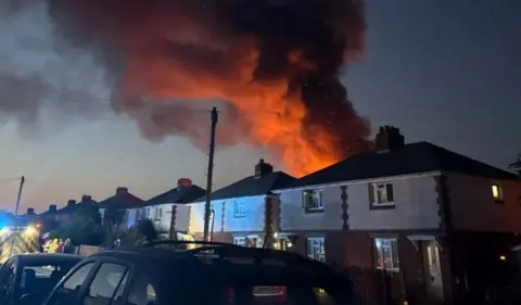 Island Echo Row of homes on a street with a fire engine seen far left. Fire and smoke is coming from behind the roof of one home.