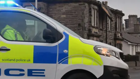 A police van in close up dominating the frame. Two officers can be seen in silhouette in the front seats. Its lights are flashing blue and there are stone houses in the background.