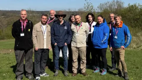 Supplied A team of 10 people, a mix of men and women, dressed in outdoor clothing standing on a grassy patch of land. There are trees in the background.