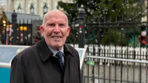Jim Rodgers stands outside the front gates of Belfast city hall. He is an older man, with short grey hair and he is smiling at the camera. He is wearing a long, dark coat, with blue and white stripes and a blue tie with white polka dots.