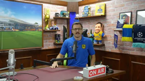 Jamie Niblock/ BBC YouTuber Spencer Carmichael-Brown his studio. There is an On Air sign, and various backdrops. He is sat in his presenting position, where he makes the videos for his channel. There are various items of football memorabilia and props around him.