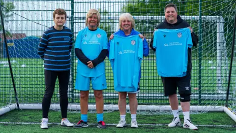 Simon Thurlow Janette Hynes, Jackie Freeman and Lisa Shepherd stood holding or wear blue football shirts in front of a goal on a football pitch. There are trees in the background. Julie Norris is wearing a stripey top.