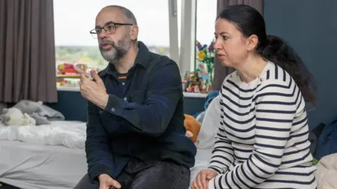 A man and a woman sit on the edge of a bed mid-conversation with someone.