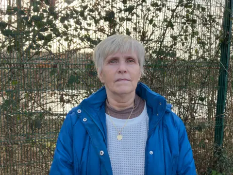 A woman stands in front of fencing. She wears a blue jacket, has short silver hair and is wearing a gold heart necklace