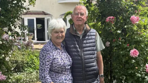 An older couple, one female and one male, David and Liz. They stand in front of an archway made of pink rose bushes. Liz has cropped grey hair and wears a floral blouse with glasses on a chain around her neck. David wears a checked shirt and dark body warmer. He wears clear glasses and has fine grey hair. Both smile.