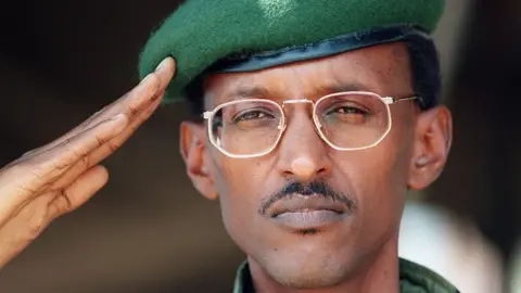 AFP via Getty Images Paul Kagame is a Black man pictured in close up. He has short black hair and a moustache and wears gold-rimmed glasses and a green military beret. The edge of a green military shirt collar can be seen at the bottom of the picture. He is saluting and looking slightly to the left of the photo. 