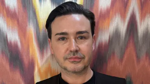 Thomas has black hair and is looking directly at the camera. He is wearing a black t-shirt and is standing in front of a a wall with colourful wallpaper. 