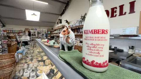 A Kimbers milk bottle is in the foreground on top of a deli counter displaying a raft of different cheeses.