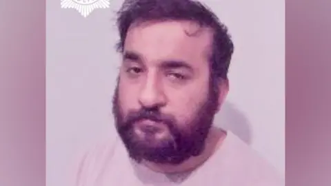 A man with dark hair and a full beard, wearing a light-coloured top against a plain background. A police crest is partially visible in the top corner of the image.