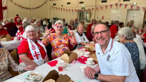 Helen Taylor/ Yaxley Parish Council A woman in a red England shirt and a white scarf, sitting next to a woman dressed in an orange-floral outfit and Andrew, a man in a white England shirt - all smiling for camera.