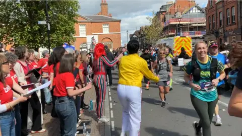 Runners grin as they pass by a Freddie Mercury lookalike in a yellow jacket and white trousers plus a person in a red and blue striped Ziggy Stardust outfit. A choir in red shirts sing on the pavement beside them. Old redbrick buildings can be seen lining the route with a police van at the side.