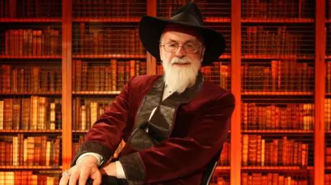 PA Media Sir Terry is wearing a read velvet jacket. His white beard is neatly groomed and he is wearing his iconic black rimmed hat. Behind him are book cases lit red.