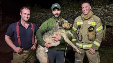 A large dog being held by a man in a green coat and a green cap.The dog is wearing a muzzle.The man has a fireman standing on either side of him.