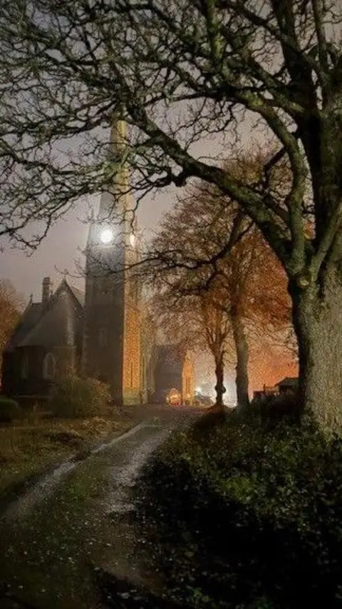 Marty Ponder A tall church with a lit clock tower stands at the end of a dim, tree-lined path.