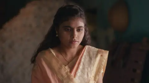 The image shows a young Indian girl clad in a traditional pale pink saree looking visibly upset. 
