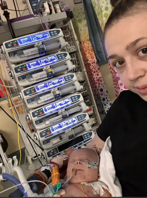 NHSBT/ PA media A woman wearing a black top is holding a baby. The baby is attached to medical equipment via tubes in his nose and mouth. Behind the woman are six IV infusion monitors - five are displaying information and the sixth is blank. 