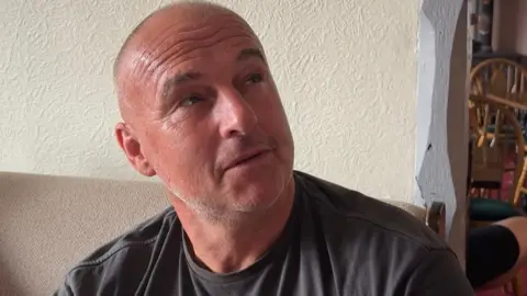 A man with a shaved head and grey stubble looks up to the right of the picture with a disdainful look on his face.