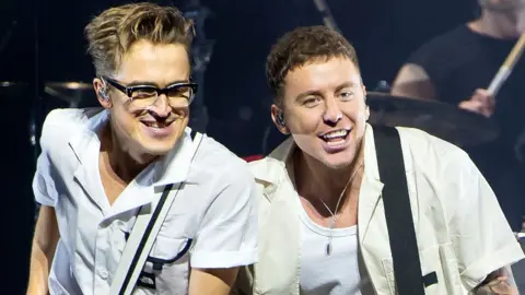 Getty Images Tom Fletcher (L) in a white short-sleeved shirt, and Danny Jones, in a cream short-sleeved shirt over a white vest, of McFly perform at The O2 Arena on November 21, 2021 in London, England