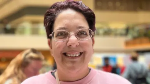 Emma Baugh/BBC A woman is smiling at the camera. She has short, dark hair and is wearing spectacles and a pink top.