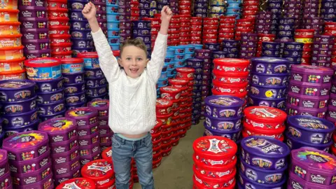 BBC Teddy wears a white jumper and jeans. He has short light brown hair. His arms are raised in triumph. He is in a warehouse full of thousands of sweet tubs, including Heroes, Quality Street, and Celebrations.