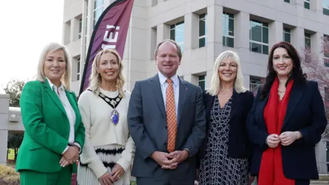 NI Chamber Four women and a man face the camera and are smiling. The man is in the middle of the picture and he is wearing a dark suit and orange tie. MIchelle O'Neill is to the left of the picture - she's wearing an emerald green trouser suit and white blouse and has shoulder length blonde hair. Beside her is a woman with long blonde curly hair who is wearing a cream coloured top and skirt with a black pattern at the top. She also wears a large medal around her neck, to signify that she's from the NI chamber. On the far right is Emma Little Pengelly wearing a dark jacket and red scarf. She has long dark hair. Beside her is another blonde haired woman wearing a dark patterned dress and dark jacket.