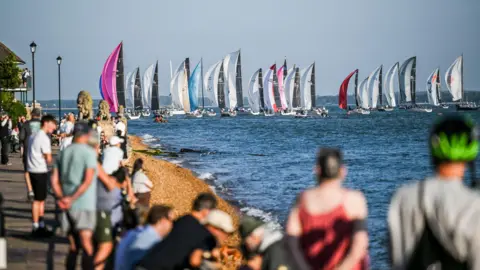 Paul Wyeth Scores of yachts lined up in the sea while spectators watch from the shoreline in the foreground