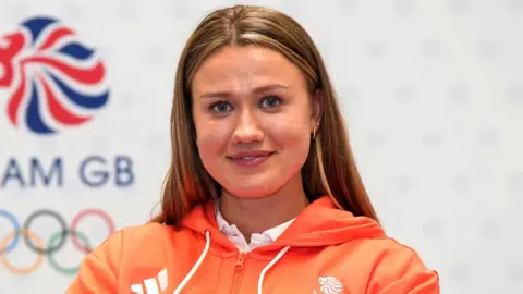 Lola Anderson has long dark blonde hair and is wearing an orange zip-up hoodie.