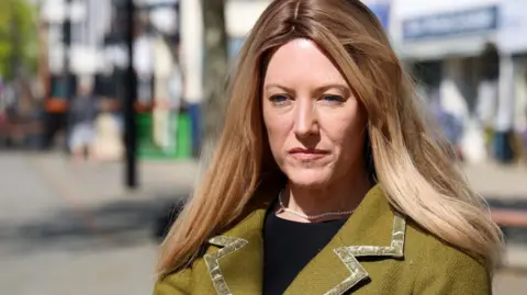 Office of Police and Crime Commissioner A head and shoulders photo of a woman with long blonde straight hair, wearing a green jacket. A blurred high street can be seen in the background.