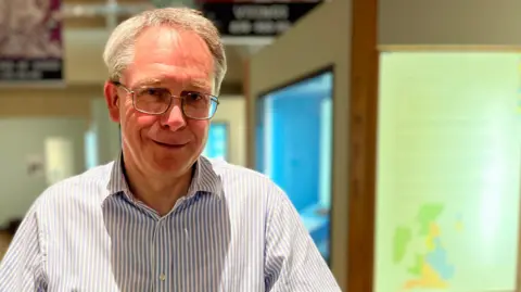 Shaun Whitmore/BBC Dr Tim Pestell standing in a gallery at Norwich castle. He is looking at the camera and smiling and is wearing a blue and white striped shirt and a pair of glasses.