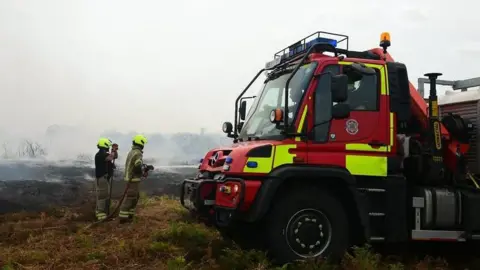 SFRS Two fire officers with hose reels jet water onto smouldering heathland. A fire service water pump is next to them. 