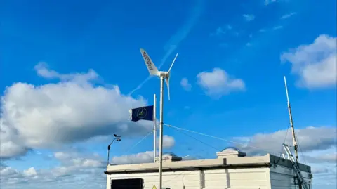 A small white wind generator pictured on a sunny but cloudy day. The white fins on the turbine say "Superwind".