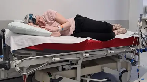 Handout Vicky Green is lying down on a a trolley in hospital. She is wearing a patterned grey and blue headscraf.