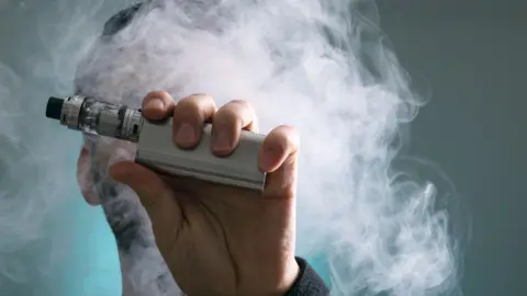 Getty Images A silver vaping device being held close to the camera. A man's face is obscured by the device and a cloud of vapour.