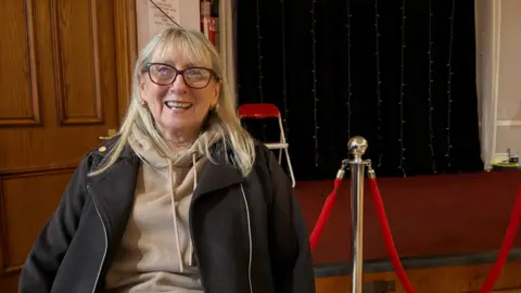 Martin Heath/BBC Yvonne Kelly with long blonde hair and brown-framed glasses smiling at the camera and wearing a beige top and dark grey cardigan. She is sitting in front of a red-carpeted stage which has a black curtain at the back and a red rope on gold poles in front.