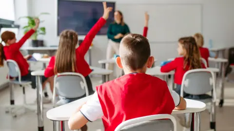 Getty Images Pupils with hands up during class. They're wearing a red jumper. A teacher is out of focus at the front of the classroom. 