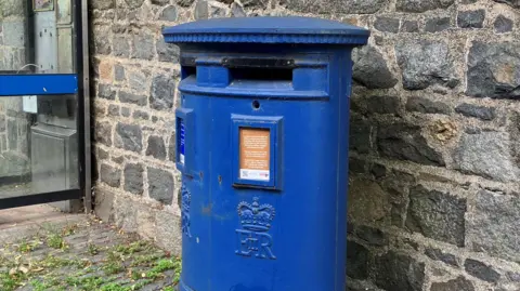 A blue double Guernsey post box in front of a Guernsey granite wall. 