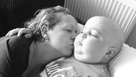 A black and white image of a woman hugging and kissing a young boy who is bald and unwell. Both of them are lying on a bed cuddling each other.